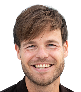 Close-up portrait of a smiling man with brown hair and light stubble, outdoors.