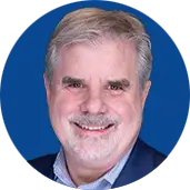 Smiling middle-aged man with gray hair and a light beard, wearing a blue blazer, against a blue circular backdrop.