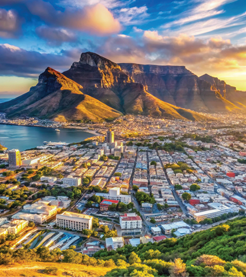 Aerial view of Cape Town at sunset with Table Mountain rising behind a coastal city and harbor.
